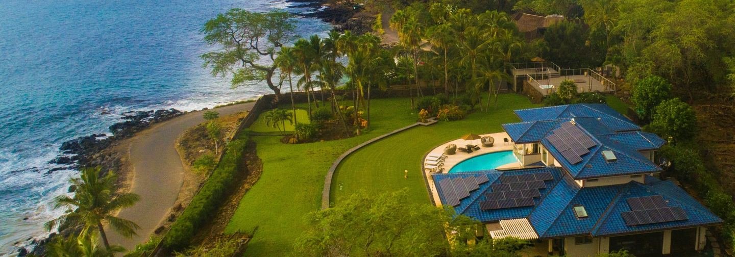 Maui coastline seen from the air: an oceanfront home with a pool and a wide lawn running down to a low seawall, a coastal road and a pocket beach of black lava and sand.