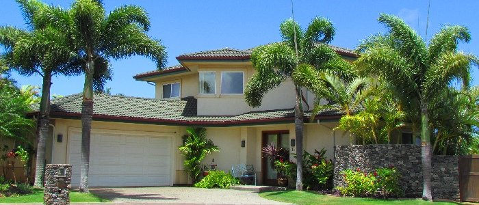 A two-story cream residential house with a two-car garage, tile roof, lava rock wall and young palms on a suburban lot.