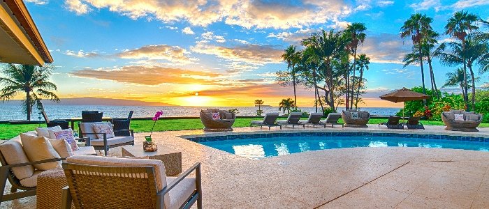 A resort-style pool and travertine deck with wicker loungers, an orchid on a table, palms and an orange sunset over the ocean.