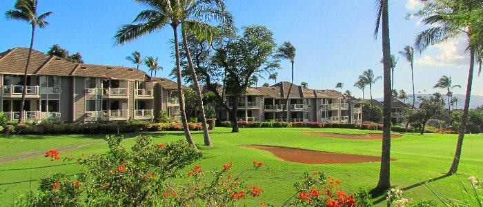 Low-rise shingle-roofed condominium buildings behind a golf fairway with a sand bunker, flowering shrubs and palms.
