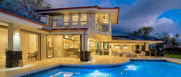 A modern two-story Maui home at twilight with a glass-walled upper room, a covered lanai and a tiled swimming pool.