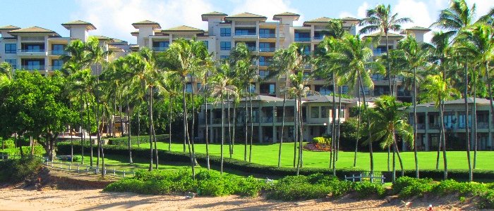 Cream stucco resort condominium buildings behind tall coconut palms, a green lawn and a sand beach.